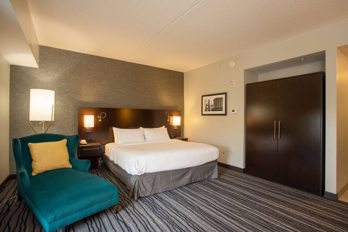 Una habitación de hotel con una cama, un sillón color turquesa, una mesita de noche con una lámpara, un armario oscuro y un cuadro en la pared.