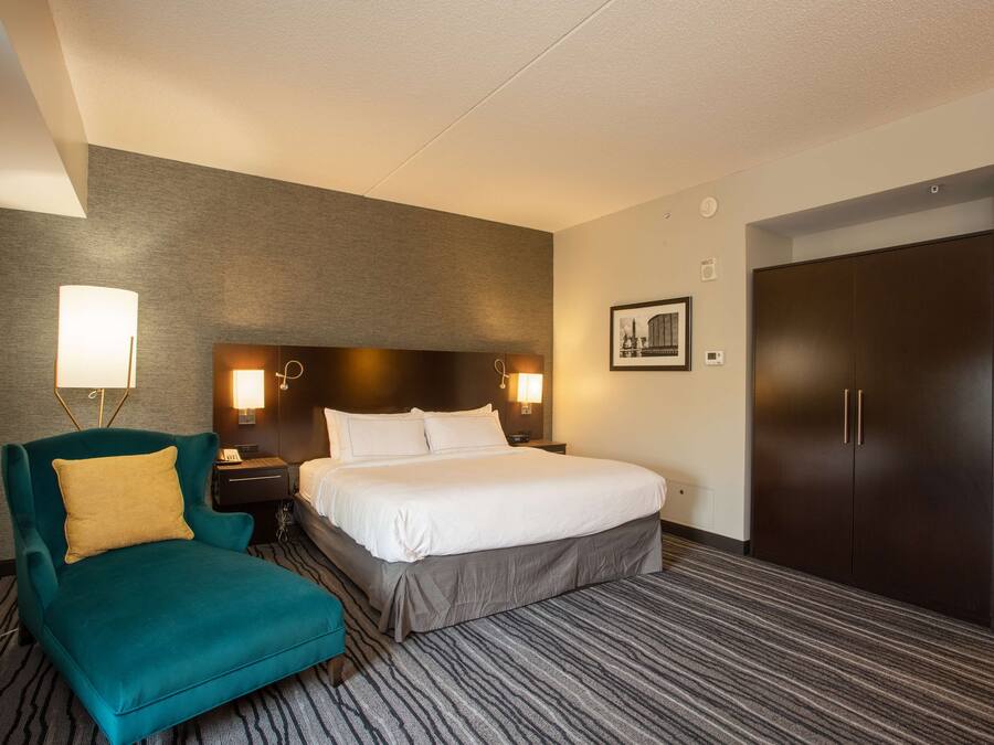 Una habitación de hotel con una cama, un sillón color turquesa, una mesita de noche con una lámpara, un armario oscuro y un cuadro en la pared.