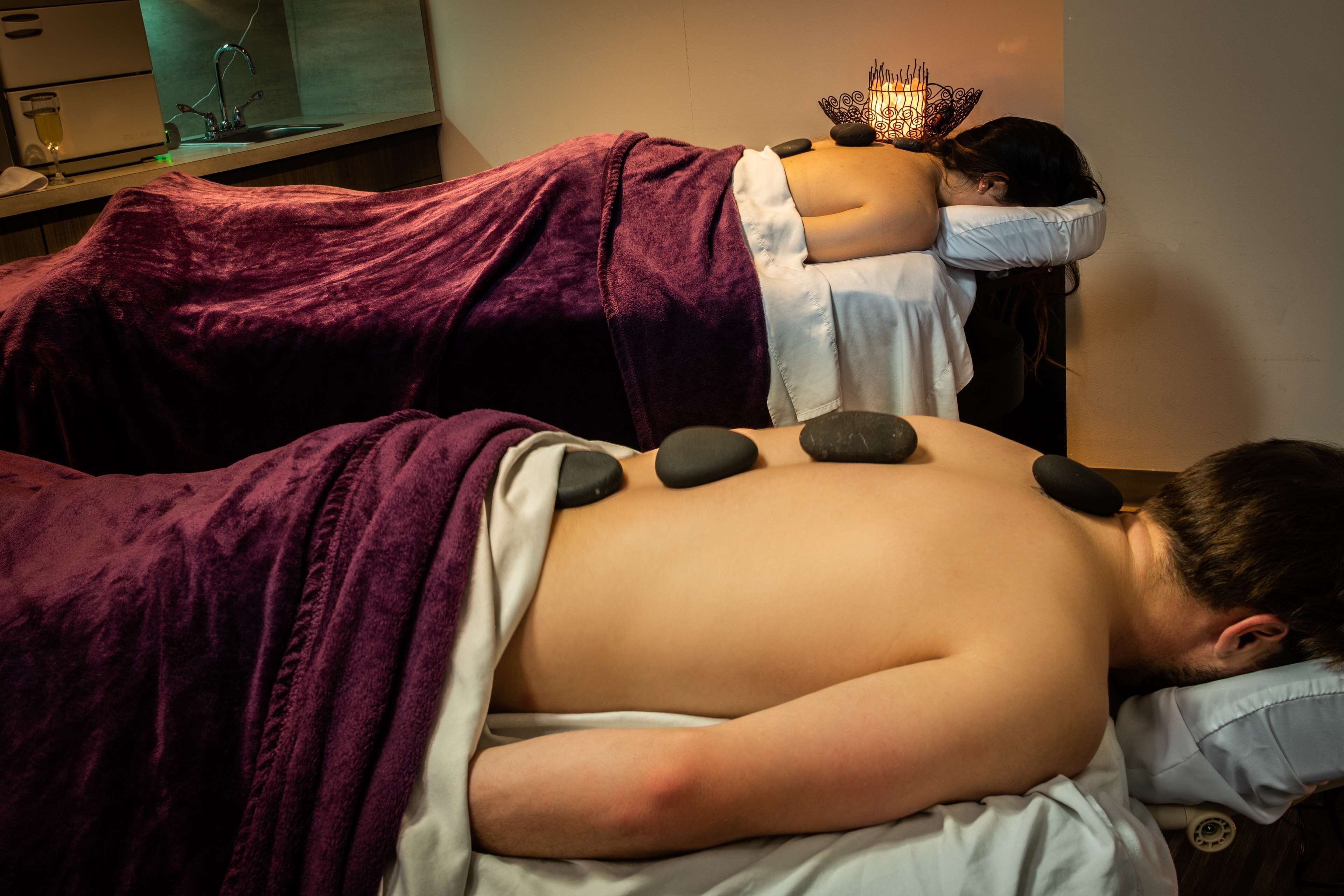 A massage therapy session with a person lying on a bed covered with a purple blanket, receiving a back massage with stones.