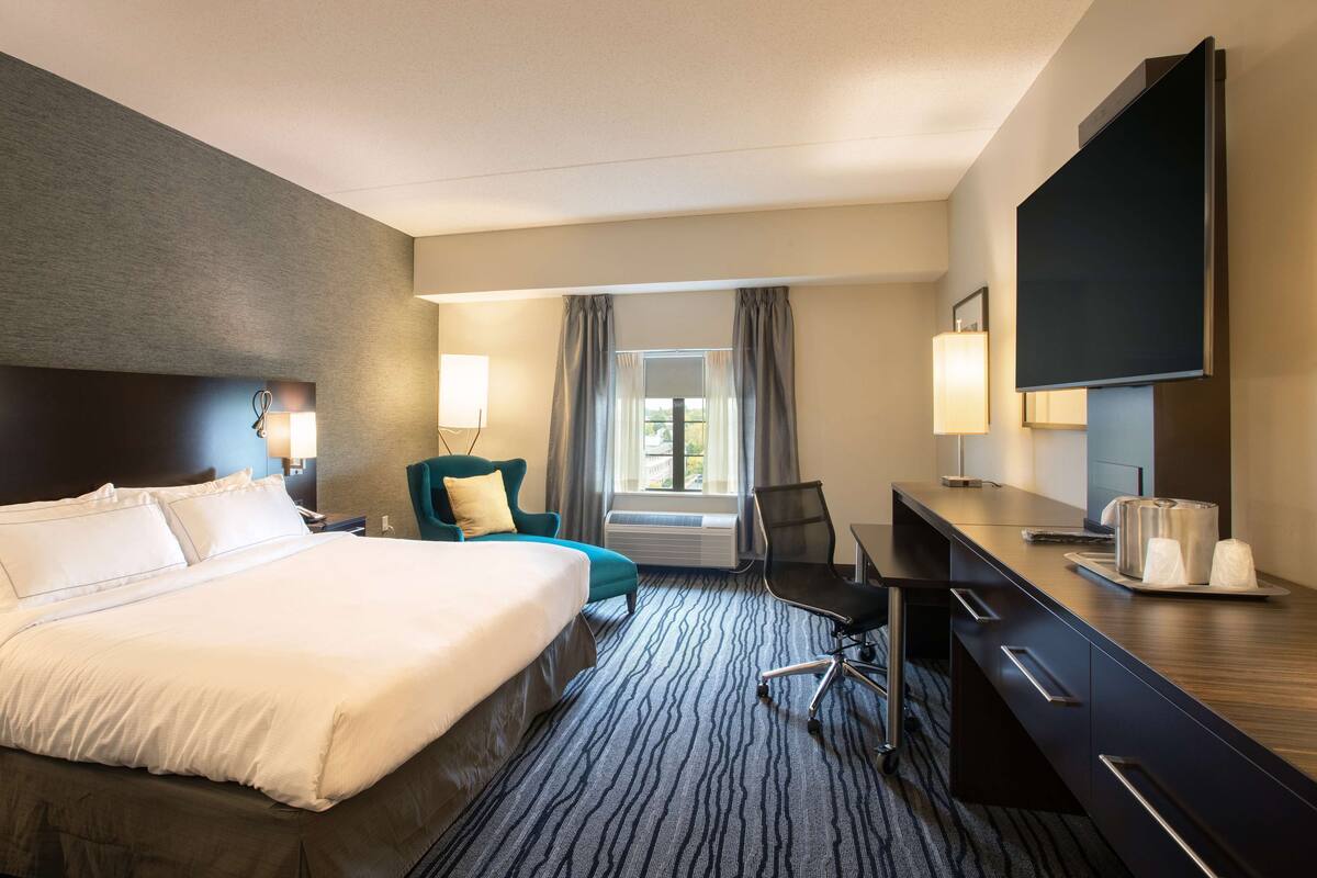 Habitación de hotel con una cama grande, un escritorio con televisor de pantalla plana, una silla, una lámpara de escritorio y una ventana con cortinas.