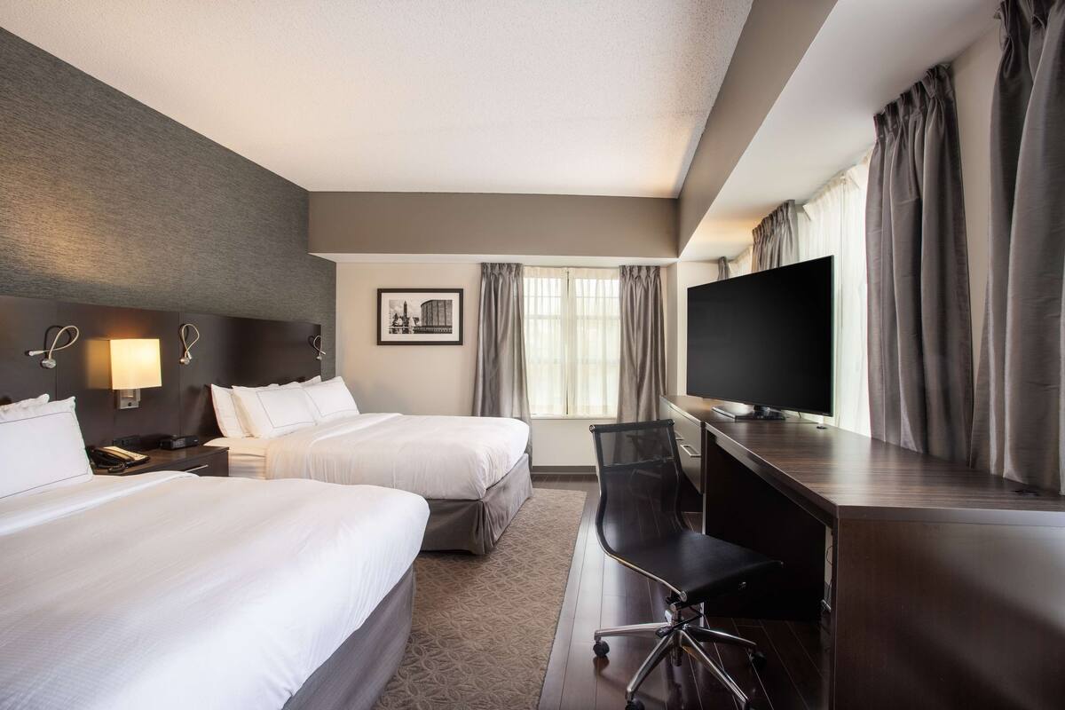 Habitación de hotel con dos camas, un escritorio, un televisor de pantalla plana, una silla y una ventana con cortinas.