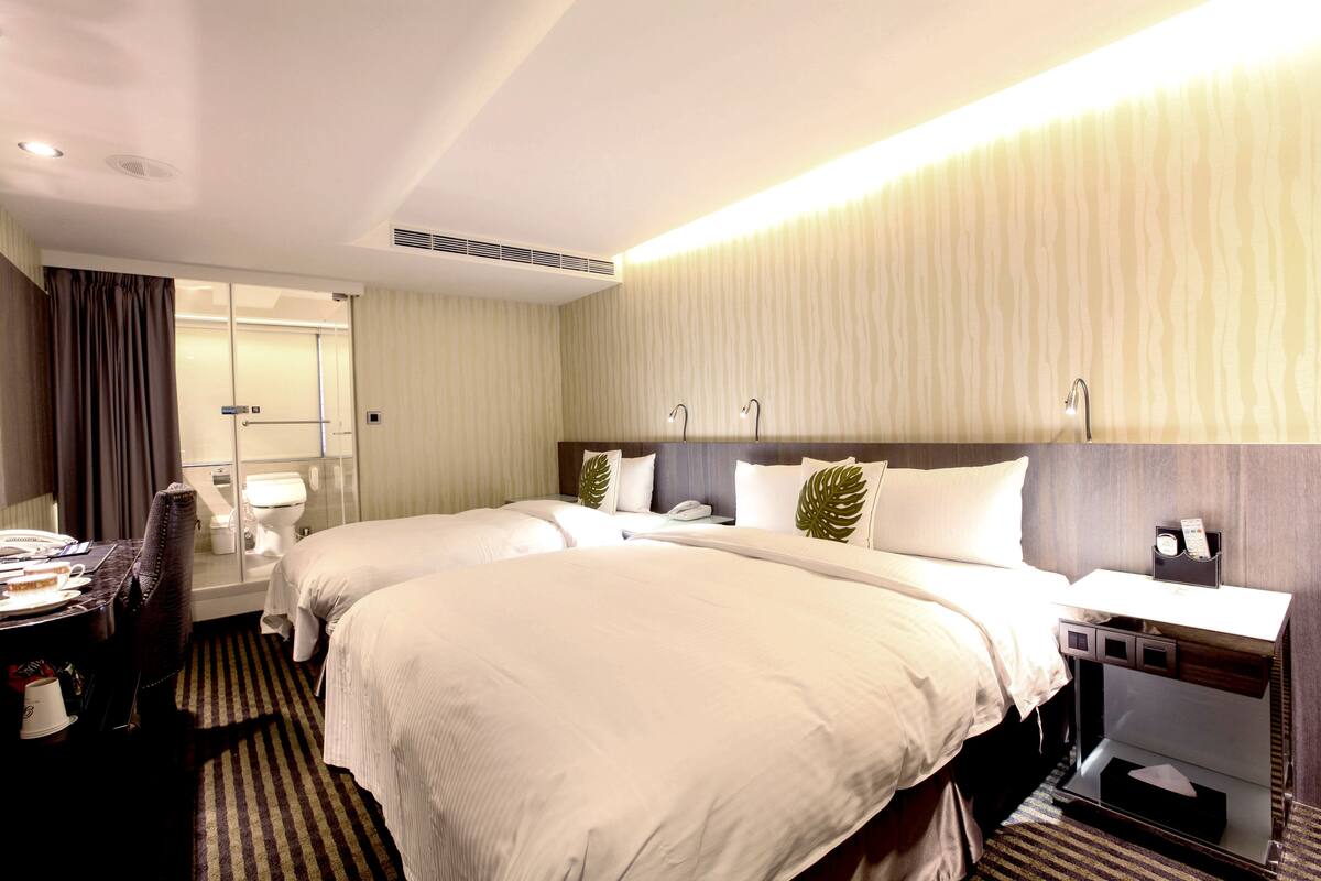 Ein Hotelzimmer mit zwei Betten, einem Schreibtisch, einem Stuhl, einem Nachttisch mit Lampe und einem durch eine Glastür sichtbaren Badezimmer.