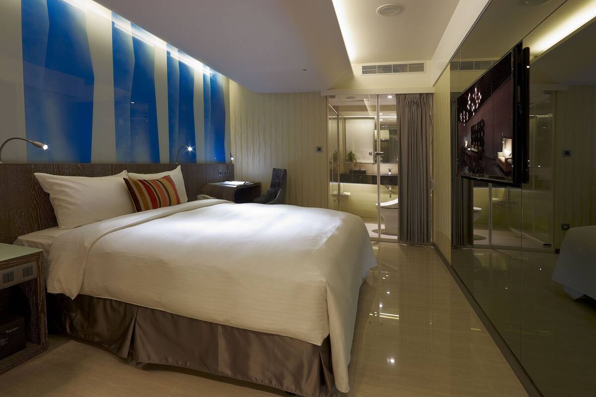 Ein modernes Hotelzimmer mit einem großen Bett, einem Schreibtisch, einem Sessel, einem Fernseher und einer Glastrennwand.