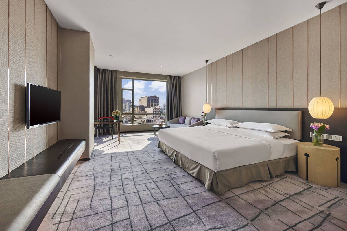 A modern hotel room with a large bed, a flat-screen TV mounted on the wall, a small desk with a chair, and a balcony with a city view.