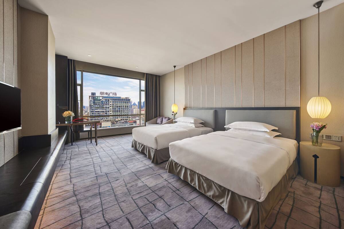 A hotel room with two beds, a TV, a desk, and a large window offering a city view.