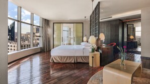 Ein modernes Hotelzimmer mit einem großen Bett, einem kleinen Beistelltisch, einer Vase mit Blumen und Blick auf die Stadt durch große Fenster.