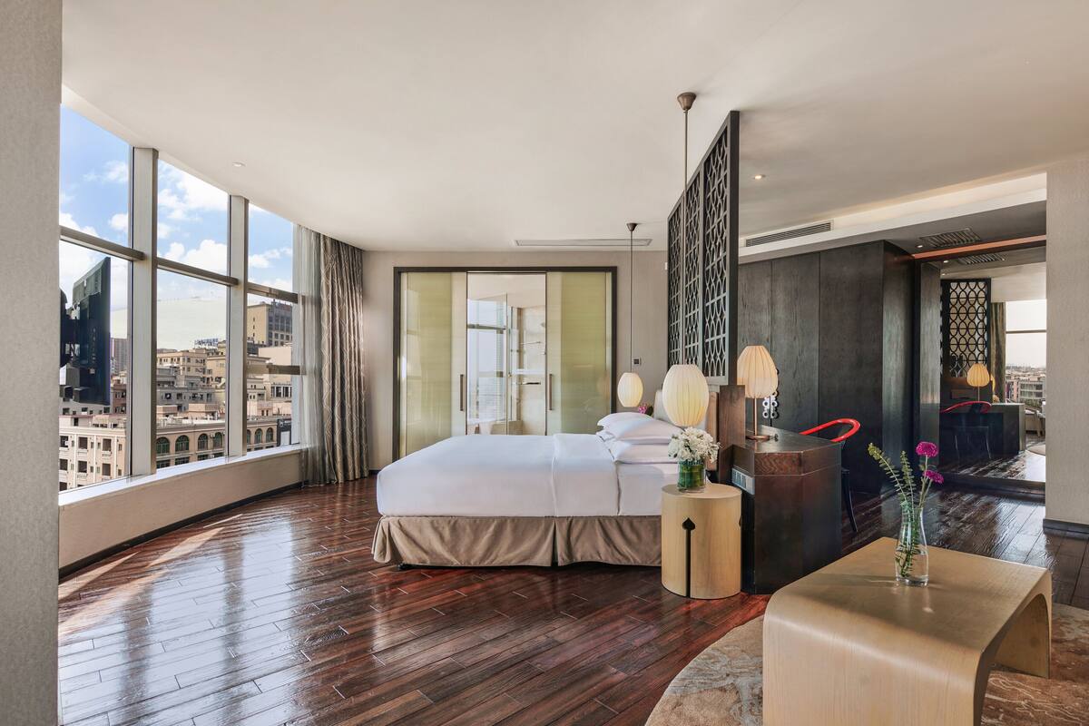 A modern hotel room with a large bed, a small side table, a vase with flowers, and a view of the city through large windows.