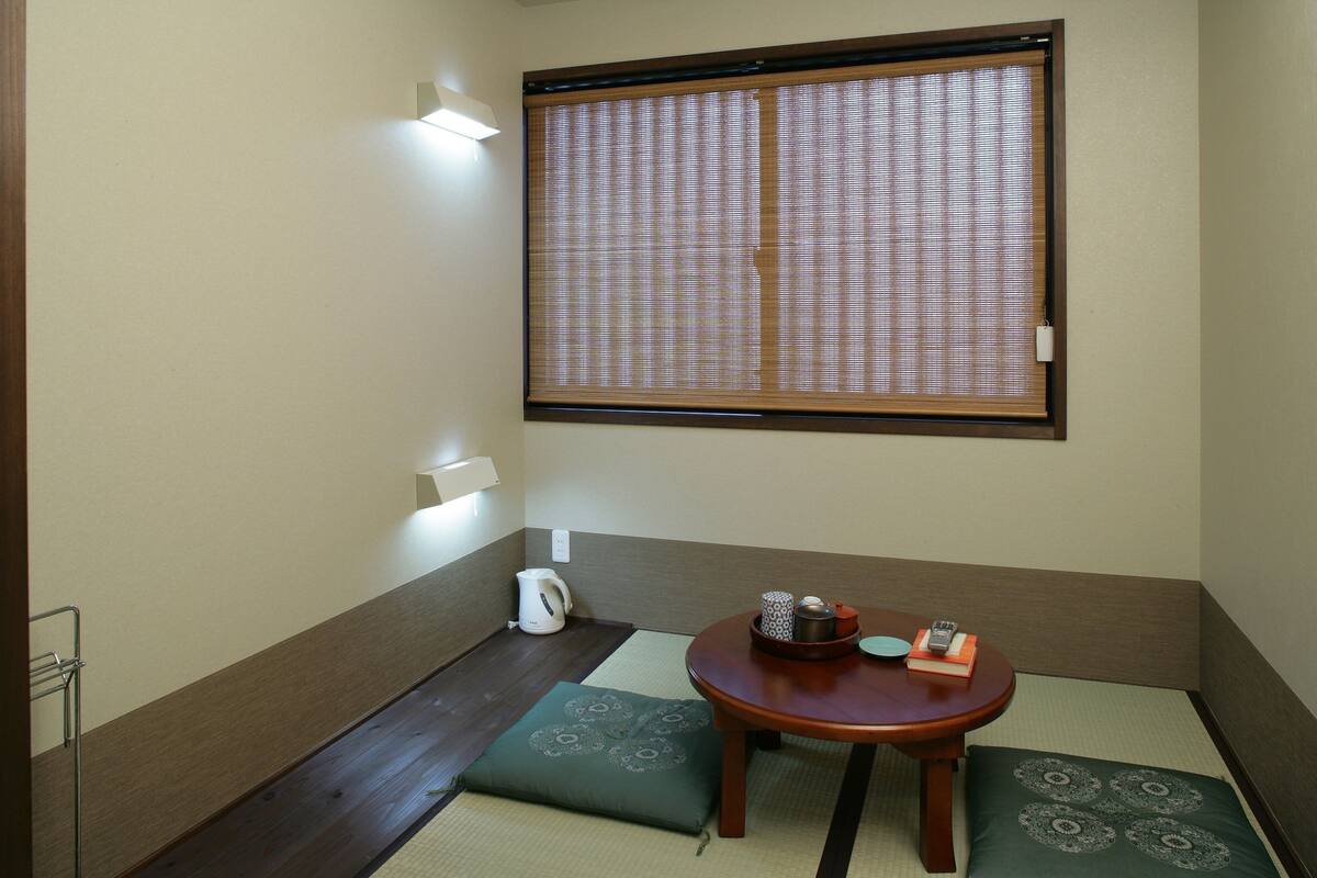 A room with tatami flooring, a low table with a cushion, a window with a blind, and a kettle on the floor.