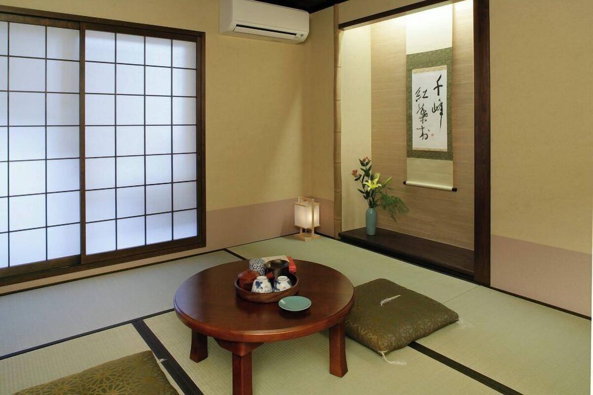 A traditional Japanese room with tatami flooring, a shoji screen, a tatami mat table with a tea set, a cushion, and a wall-mounted scroll.