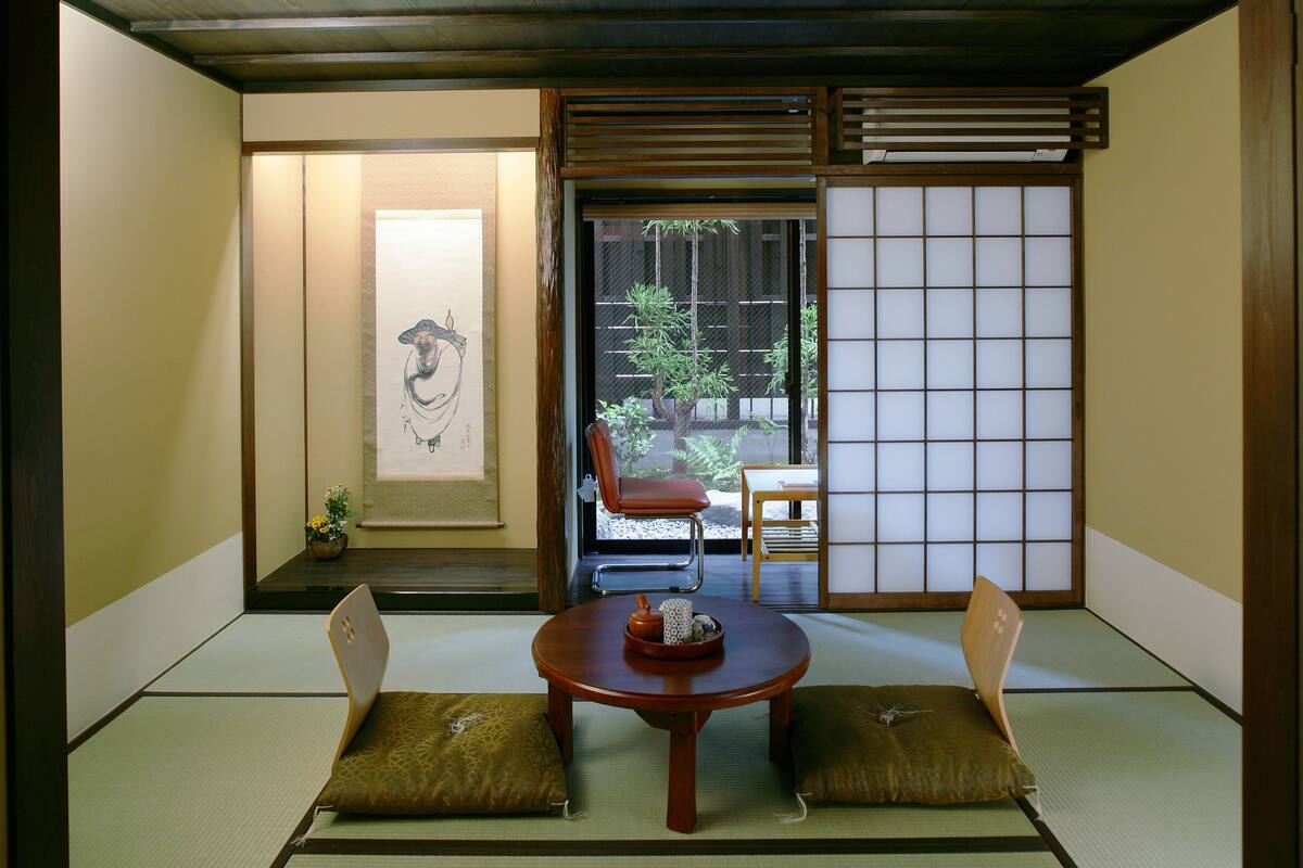 A traditional Japanese room with tatami flooring, a low wooden table, two cushions, a sliding door, and a view of the outside.