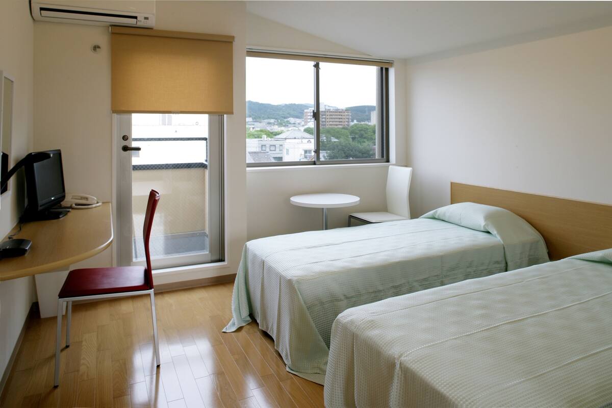 A hotel room with two beds, a desk with a chair, a television, and a window with a view of buildings.