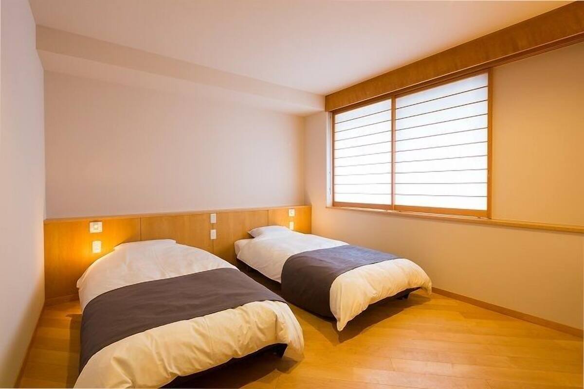 A small, single-room accommodation with two beds, a wooden headboard, a window with blinds, and a light switch on the wall.