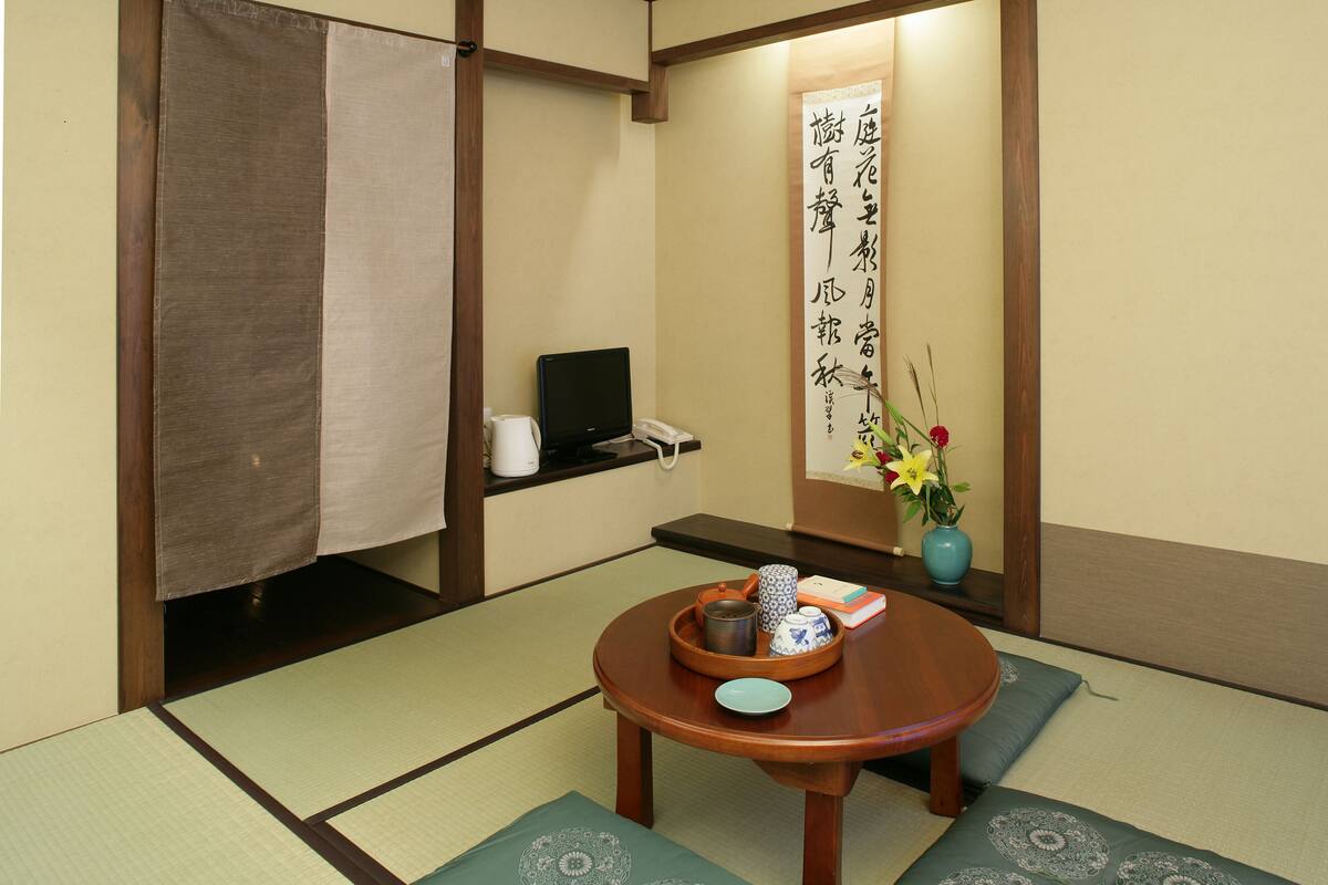 A traditional Japanese room with tatami flooring, a tatami mat table, a small wooden table, a vase with flowers, and a calligraphy scroll on the wall.