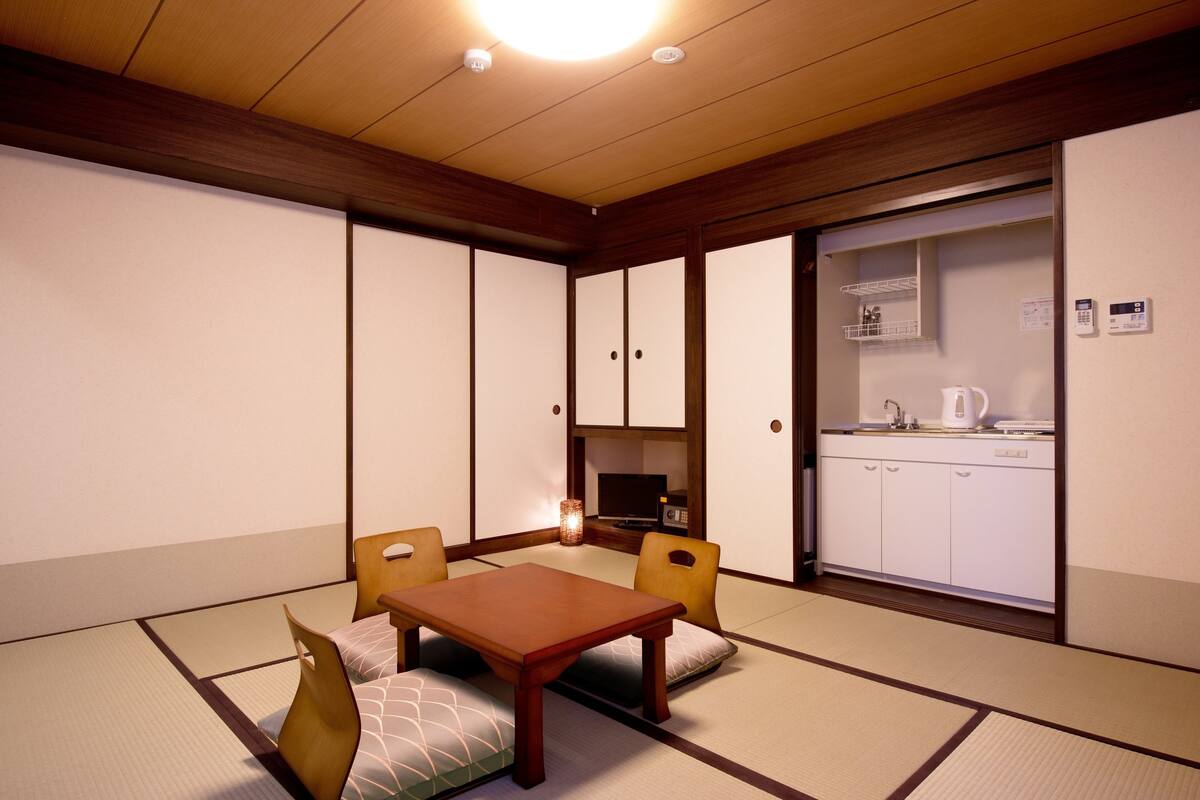 A traditional Japanese room with tatami flooring, a low wooden table, and a kitchen area with white cabinets and a coffee maker.