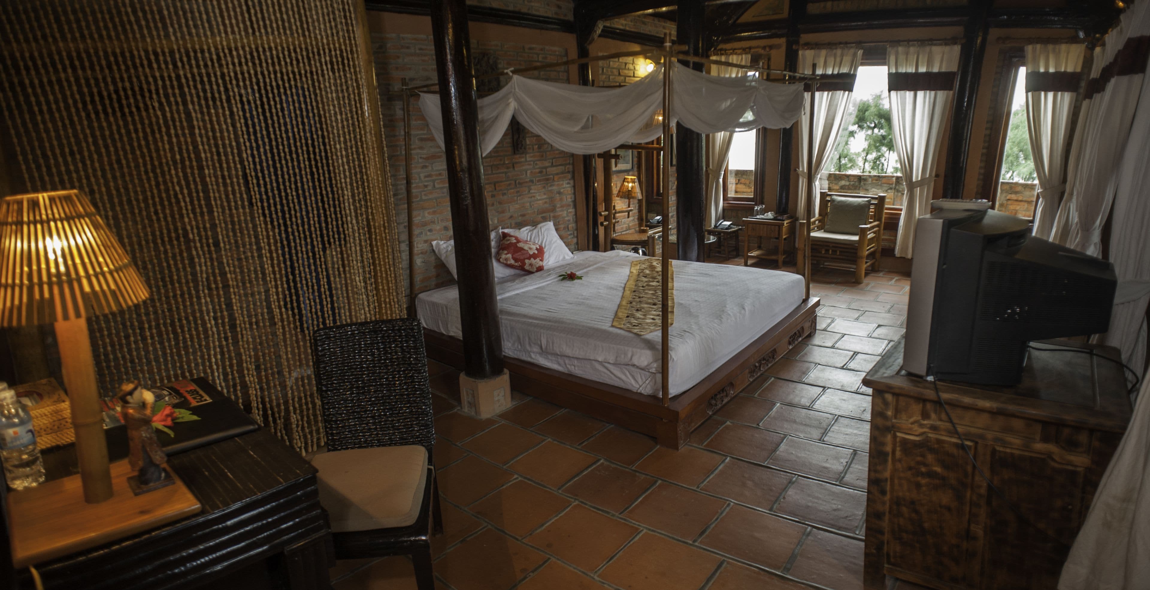 A bedroom with a four-poster bed, a wooden desk, a chair, a television, and a balcony with a view.