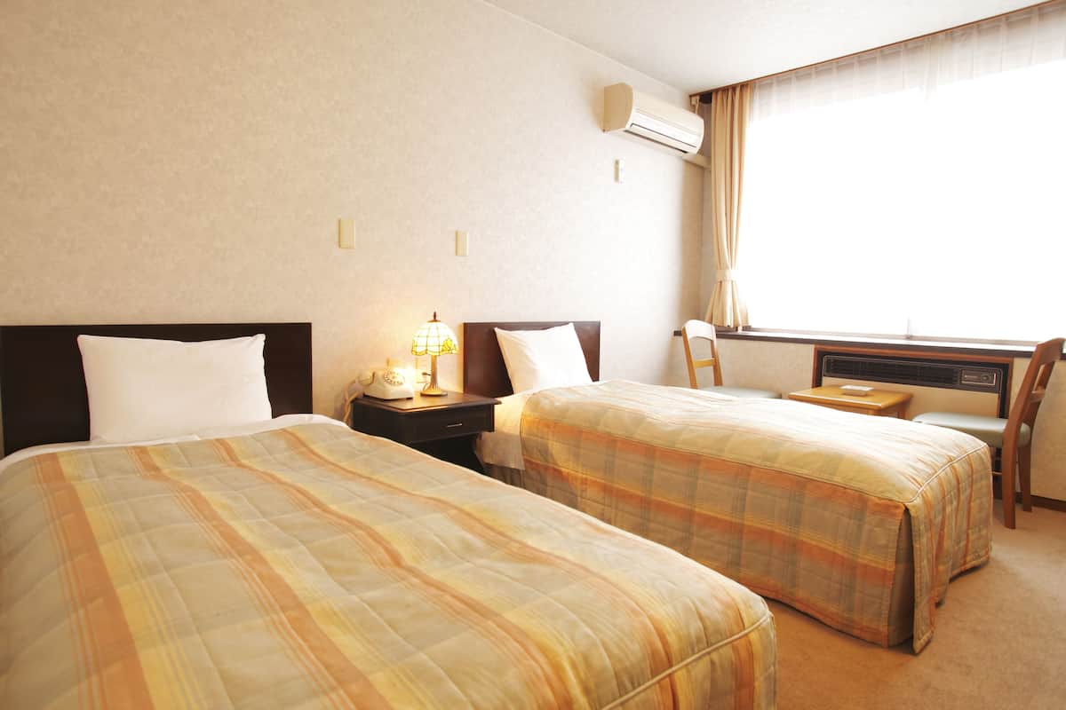 A hotel room with two beds, a nightstand with a lamp, a chair, a desk, and a window with curtains.