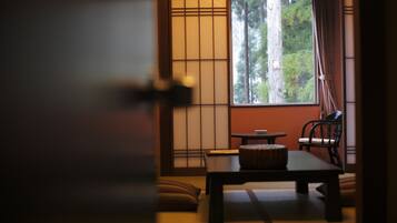 A traditional Japanese room with tatami flooring, a low table, a chair, and a window with a view of trees.