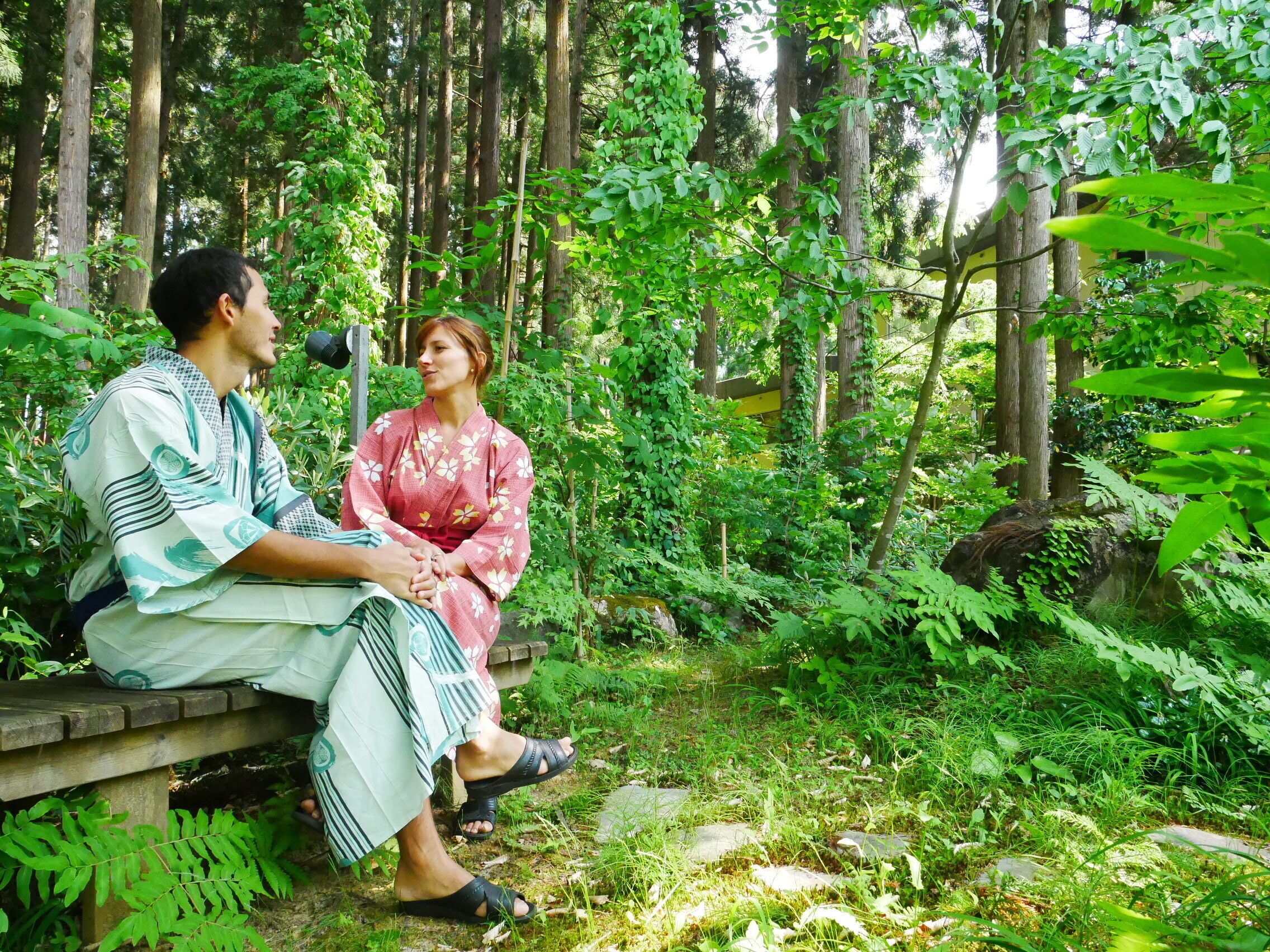 A couple sitting on a wooden bench in a forested area with dense greenery and tall trees.