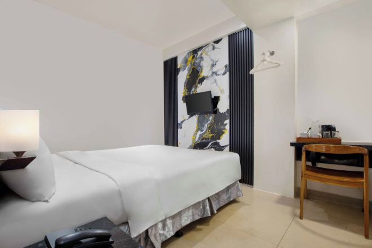 A hotel room with a bed, a desk, a chair, a lamp, and a TV mounted on the wall.