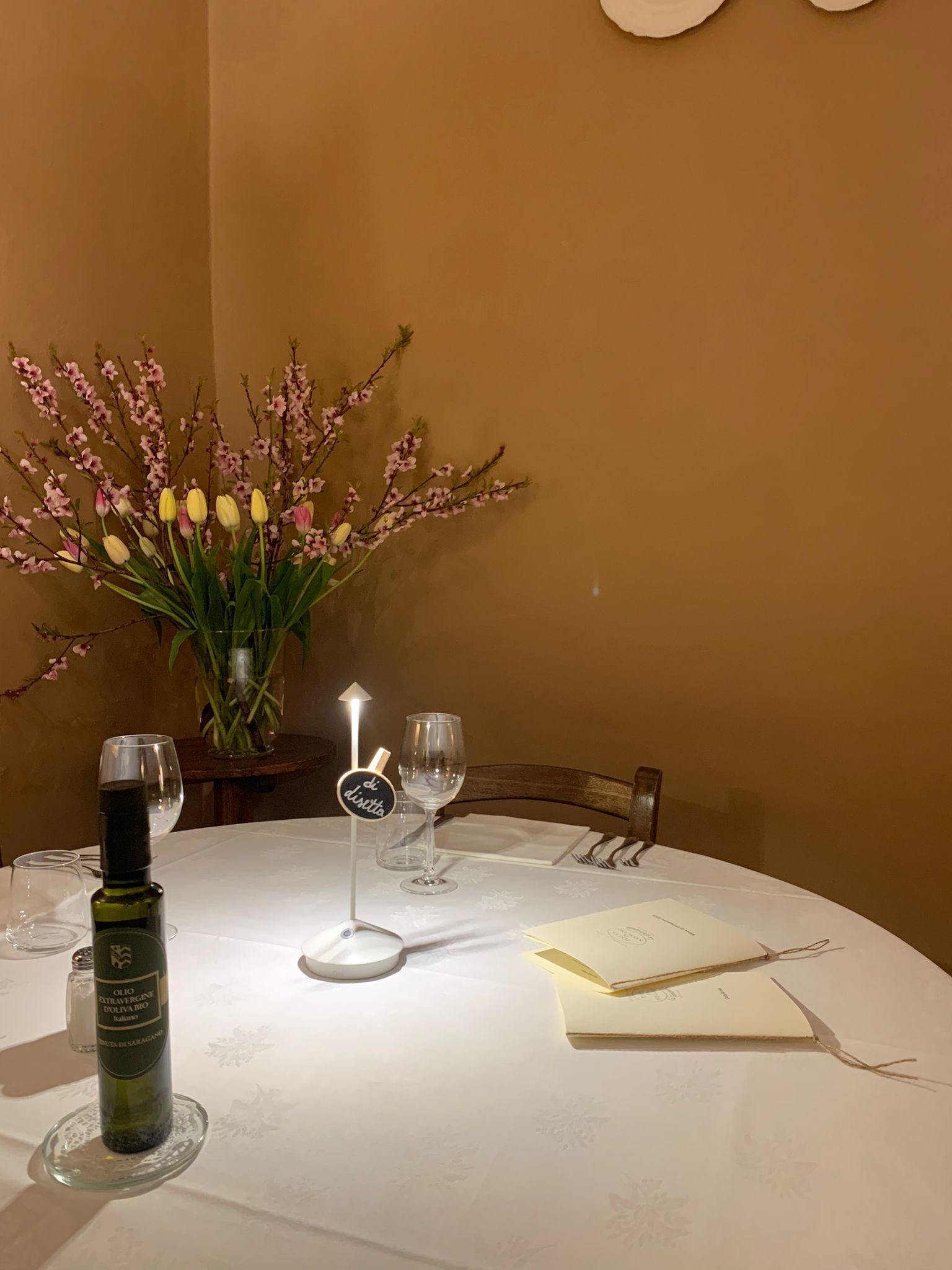 Een eettafel gedekt met een fles olijfolie, twee wijnglazen, een menukaart en een bloemstuk.