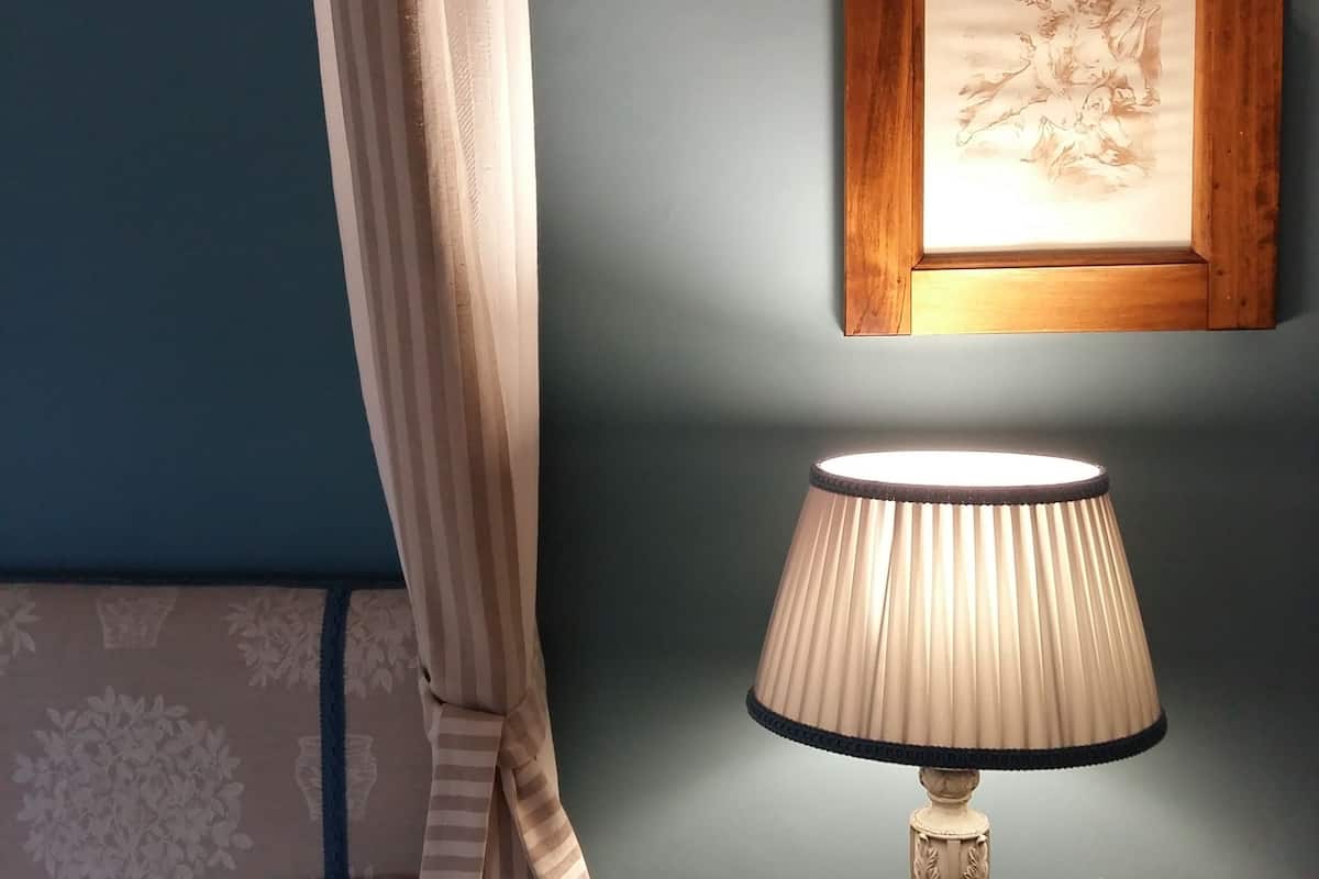 A bedroom with a bed, a bedside table with a lamp, a wall-mounted picture frame, and a curtain.