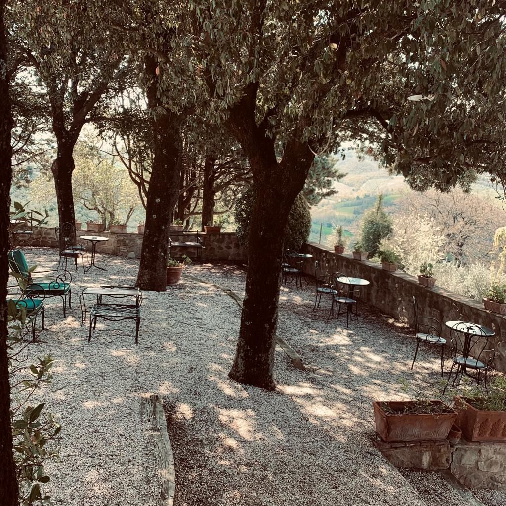 Un espacio al aire libre con mesas y sillas, rodeado de árboles y un muro de piedra.