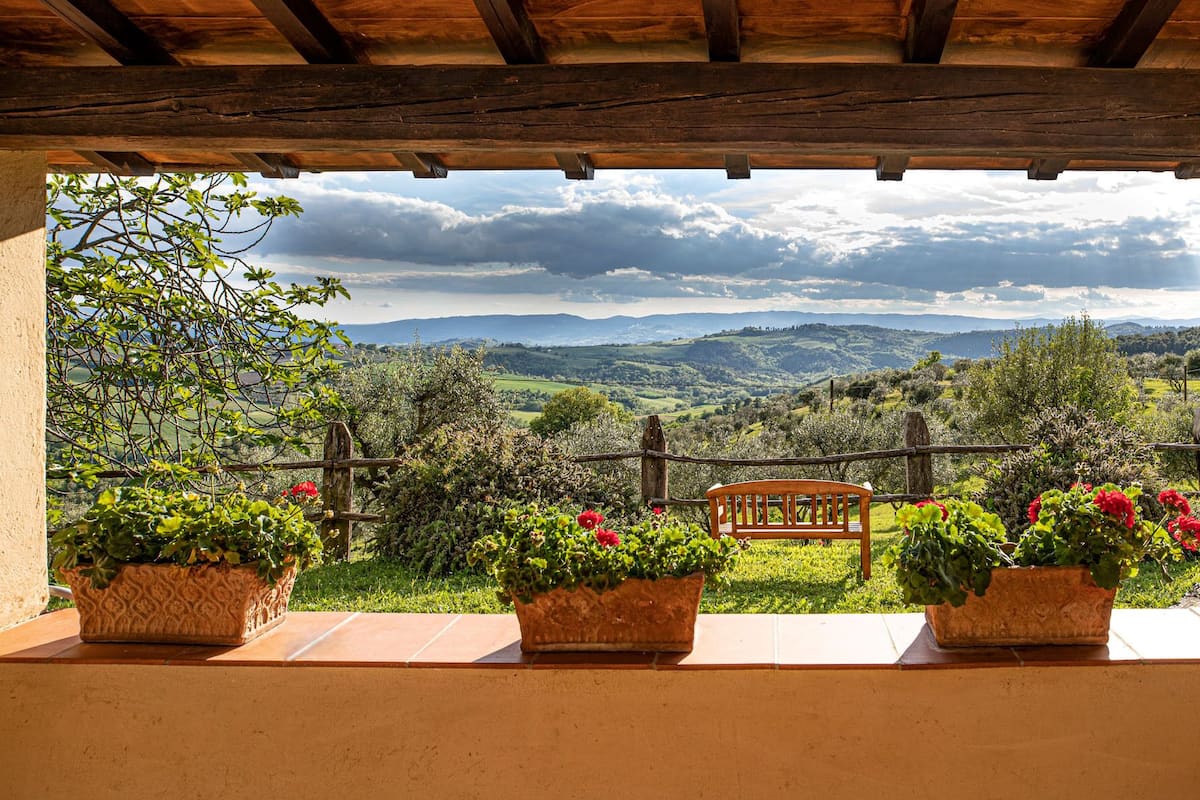 A covered patio with a wooden bench and potted plants, overlooking a scenic landscape with hills and a cloudy sky.