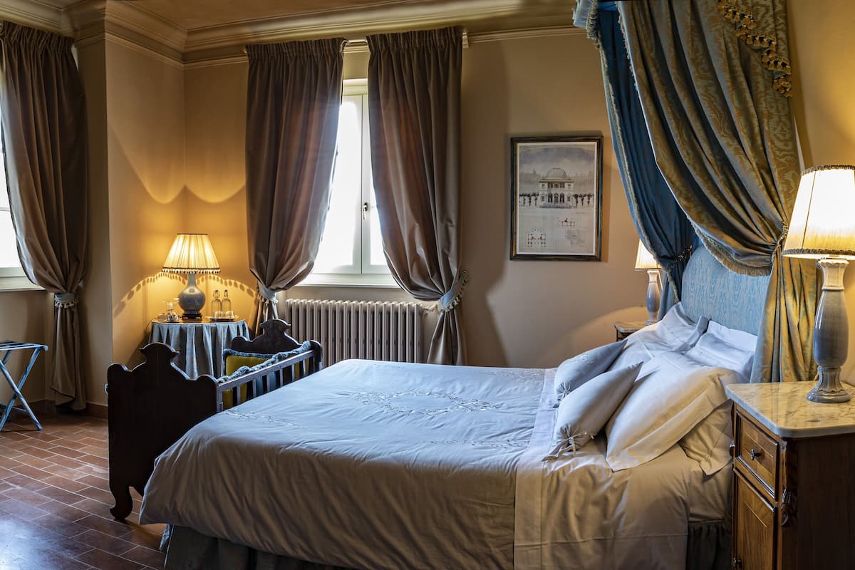 A bedroom with a large bed, two bedside lamps, a small table with a lamp, a radiator, and a window with curtains.