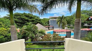 Ein Balkon mit Blick auf einen Swimmingpool, Palmen und ein Gebäude mit rotem Dach.