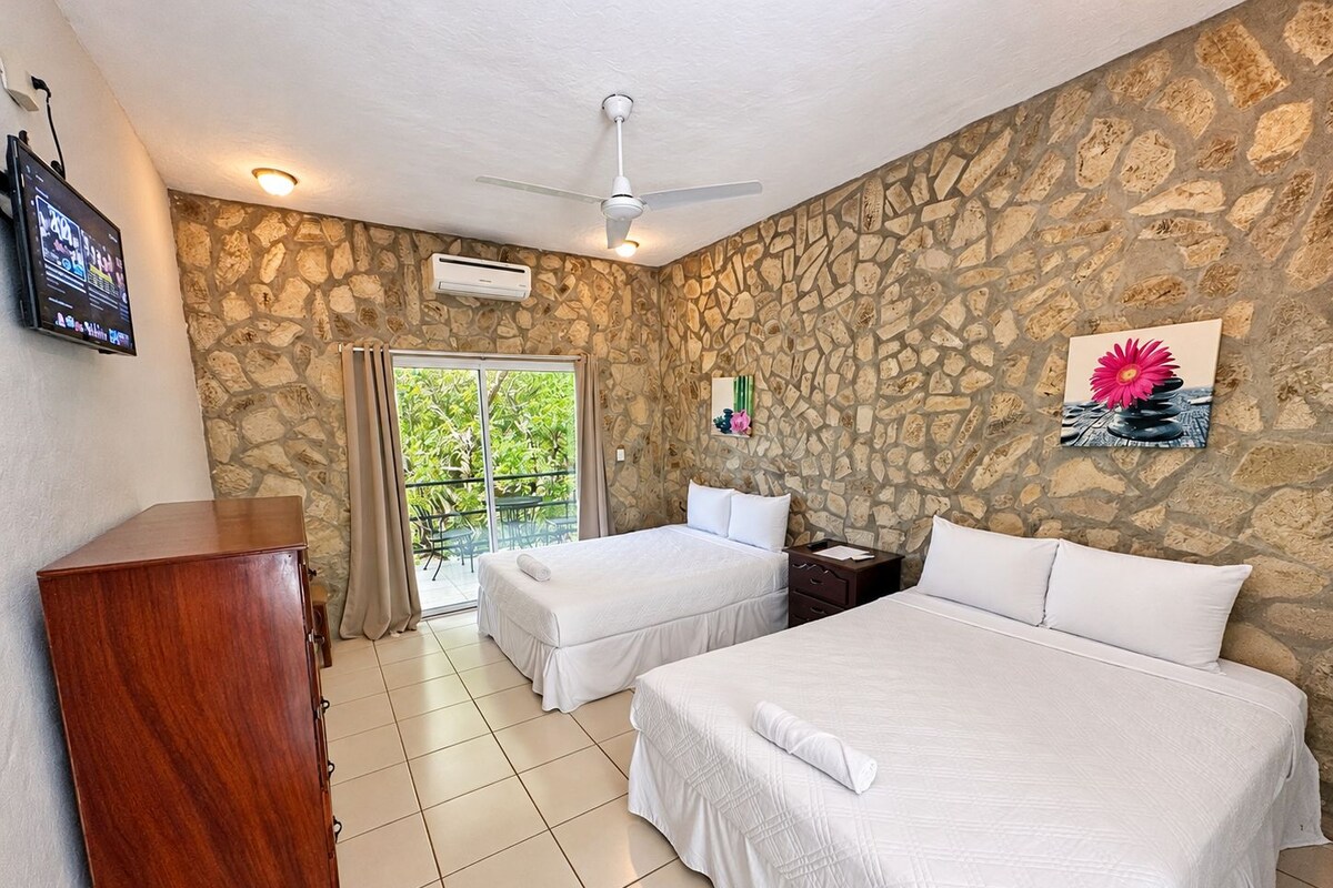 A hotel room with a stone wall, a bed, a desk, a television, and a balcony with a view of greenery.