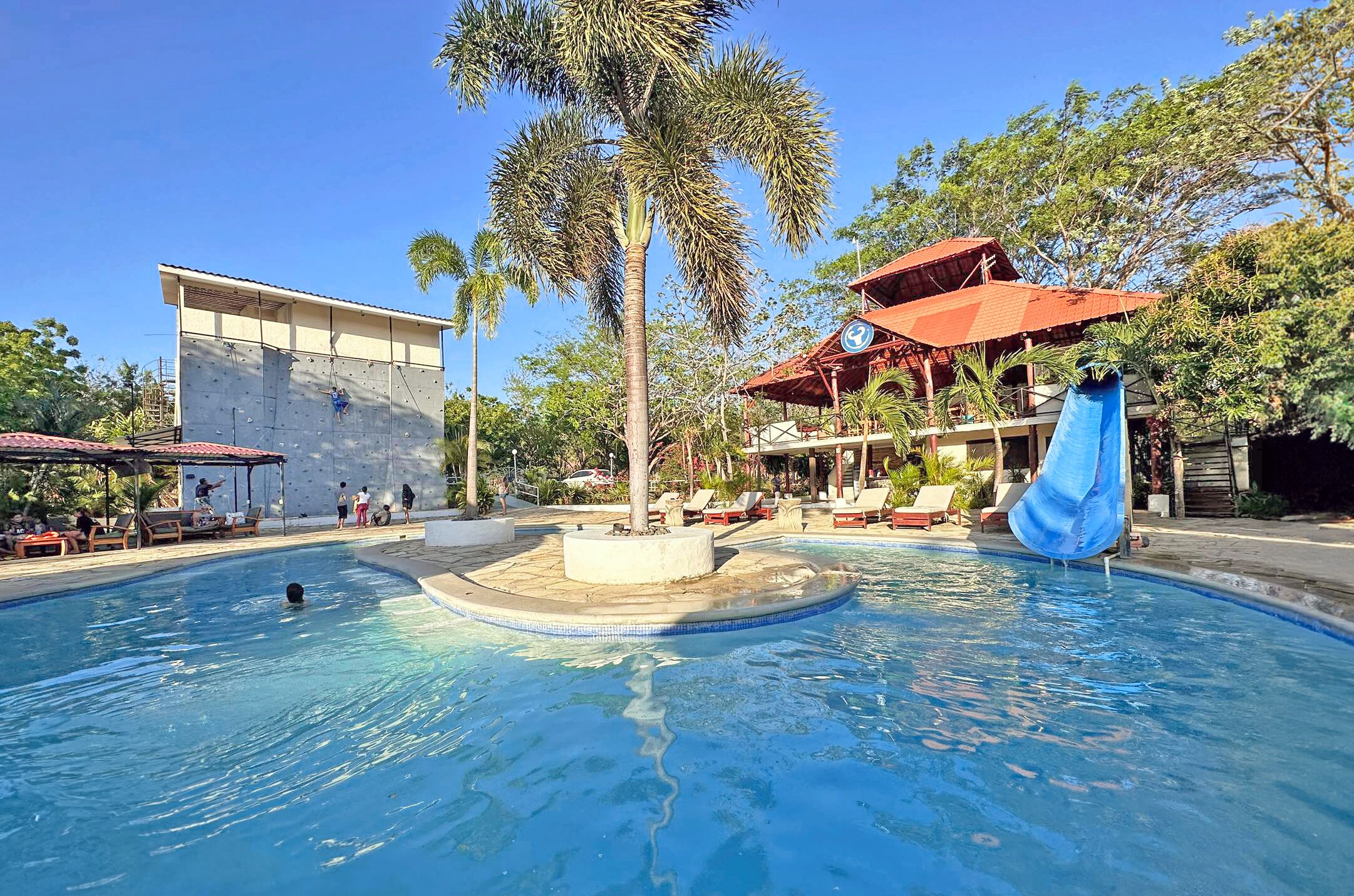 Piscina com escorregador, palmeiras e um edifício com telhado vermelho.