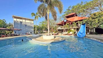 Piscina com escorregador, palmeiras e um edifício com telhado vermelho.