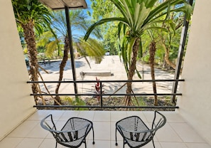A balcony with two chairs, a view of a sandy area with palm trees, and a bench.