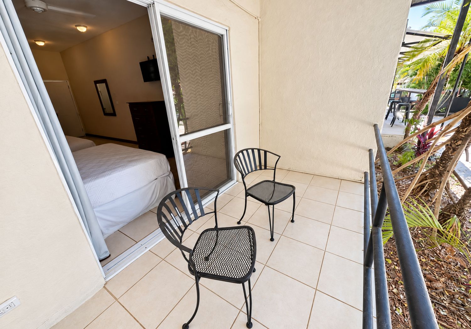 Un balcon avec un lit, deux chaises et une vue sur des palmiers.