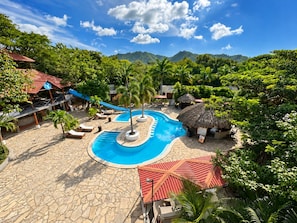 A resort pool area with a central pool, lounge chairs, and a thatched-roof hut.