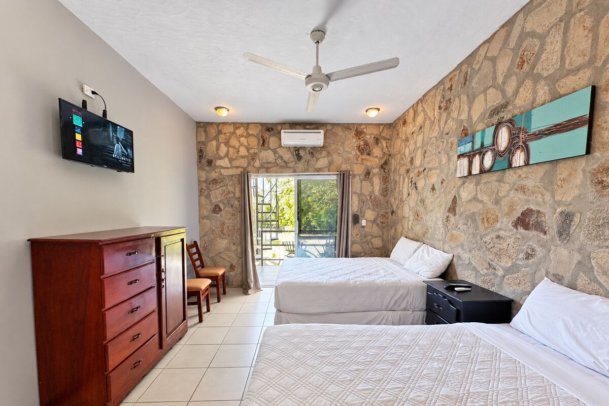 A hotel room with a stone wall, a ceiling fan, a TV, a wooden dresser, two beds, and a balcony with a view.