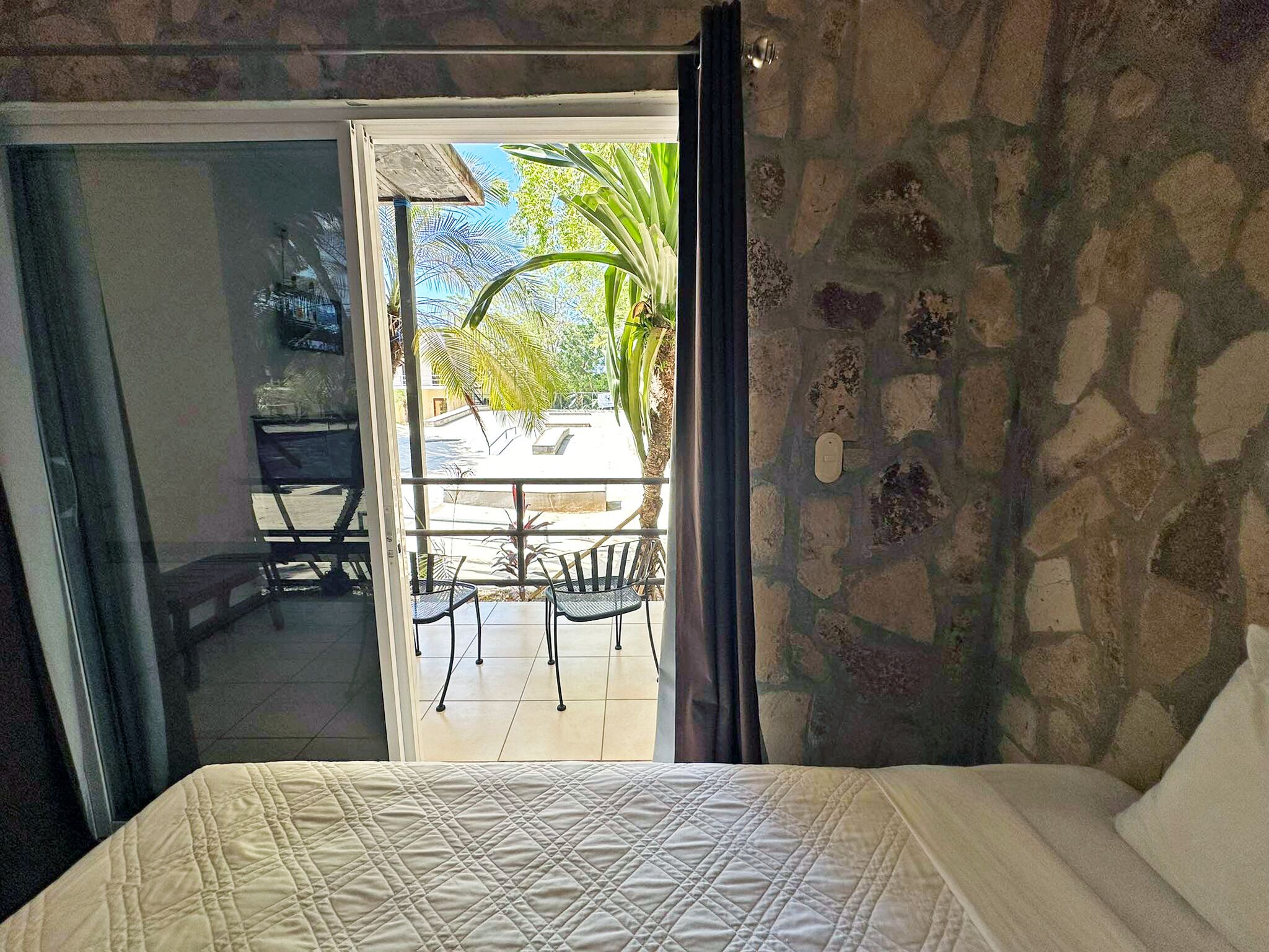 A bedroom with a bed, a sliding glass door leading to an outdoor area with a table and chairs, and a stone wall.