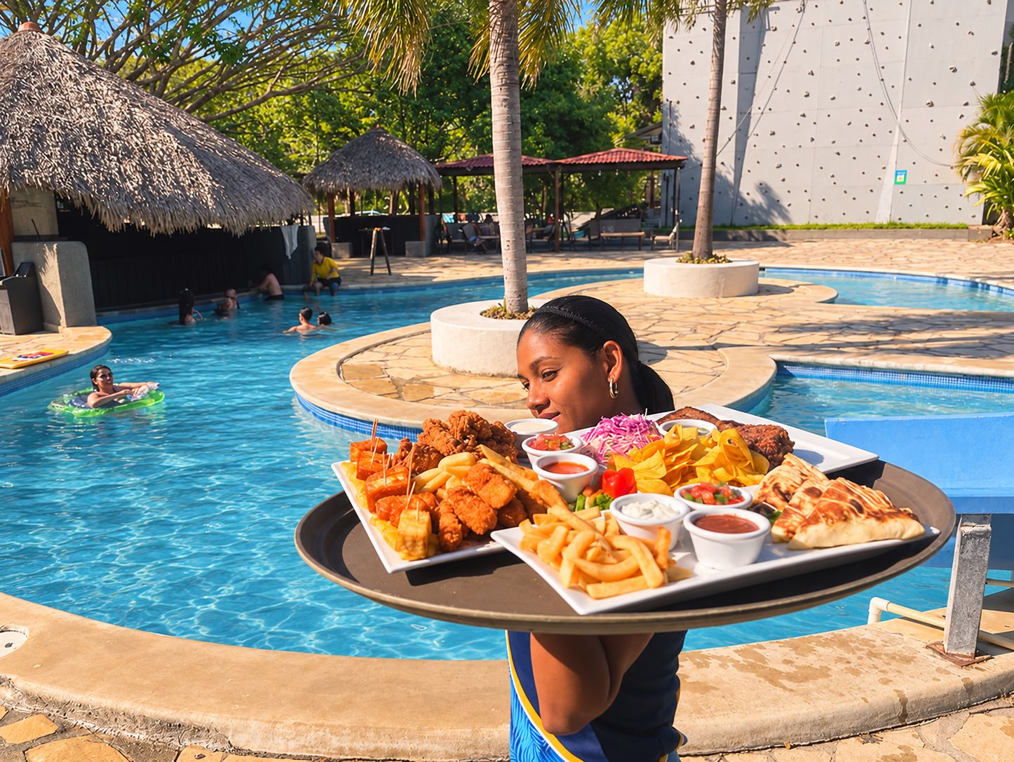 Un espace repas aménagé au bord de la piscine, avec un plateau de nourriture comprenant des plats frits et des chips, et une piscine où des personnes nagent et se détendent.