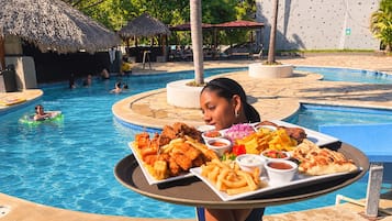 Un espace repas aménagé au bord de la piscine, avec un plateau de nourriture comprenant des plats frits et des chips, et une piscine où des personnes nagent et se détendent.