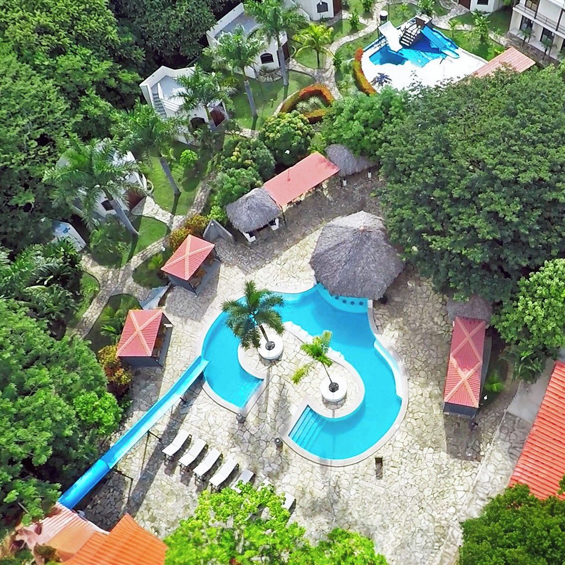 An aerial view of a resort with multiple swimming pools, palm trees, and thatched-roof structures.