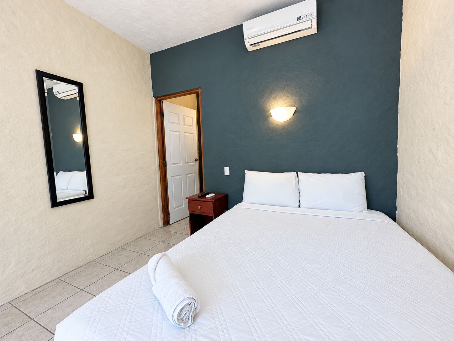 A hotel room with a bed, a nightstand, a mirror, a door, and an air conditioning unit.