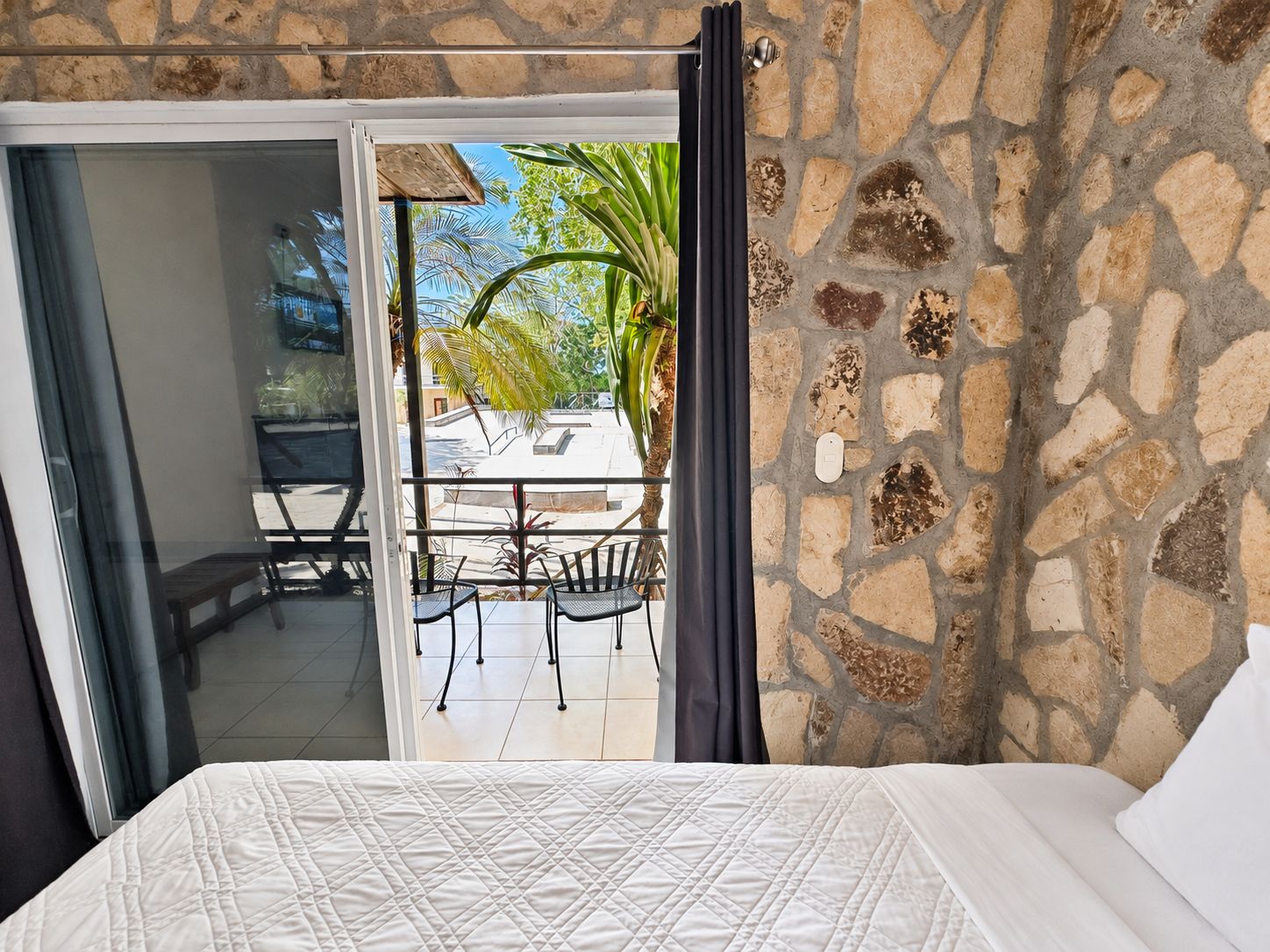 A bedroom with a bed, a view of an outdoor balcony with chairs and a table, and a stone wall.