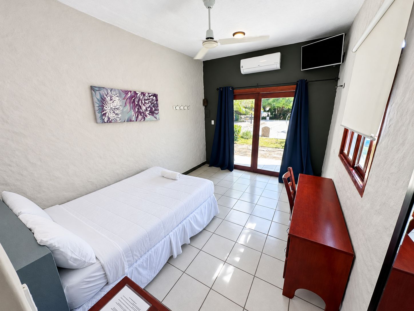 A hotel room with a bed, a desk, a chair, a ceiling fan, an air conditioning unit, a TV, and a window with curtains.