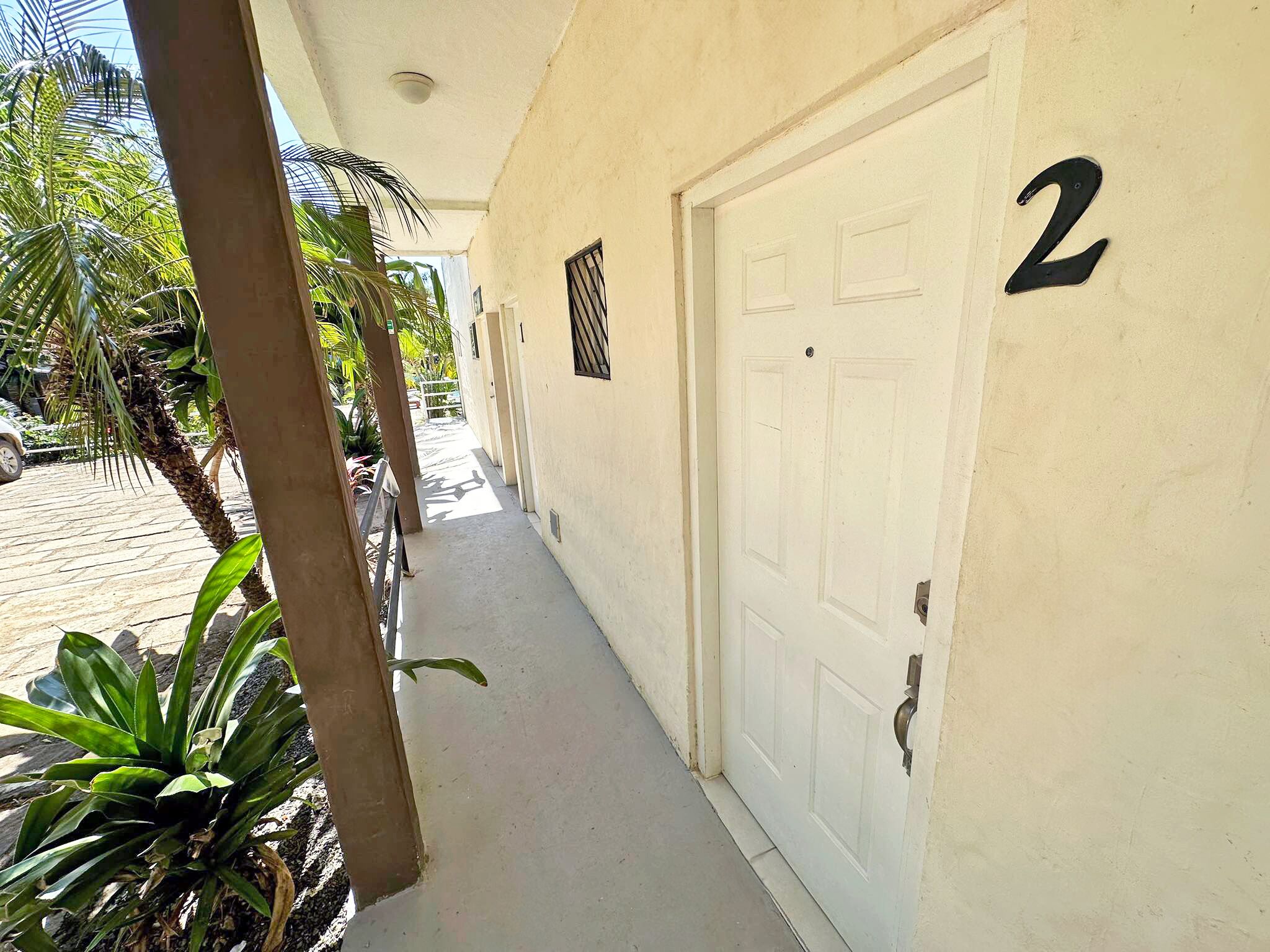 Un pasillo con una puerta blanca numerada 2, una reja de metal y una palmera.