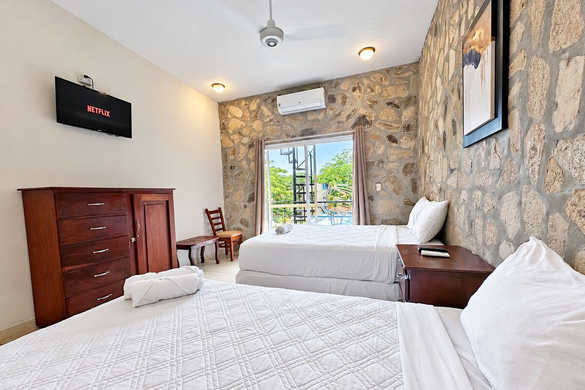 A hotel room with two beds, a wooden wardrobe, a TV, and a stone wall.
