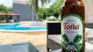 Eine Flasche Tona Bier mit einem Etikett, das Palmen und Berge zeigt, auf einem Tisch in der Nähe eines Poolbereichs mit Blick auf ein Gebäude und Grünflächen.