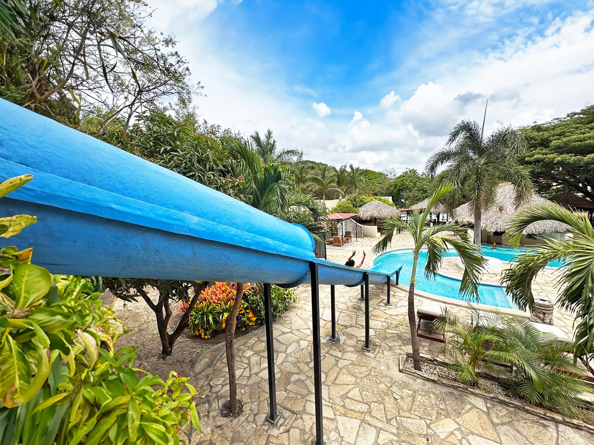 A pool area with a blue slide, surrounded by tropical plants and a stone pathway.