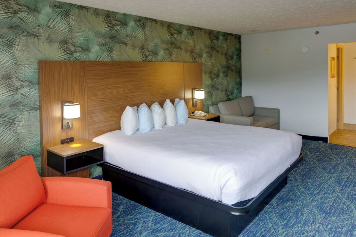 A hotel room with a large bed, two bedside tables with lamps, a wooden headboard, a red chair, and a door to the right.