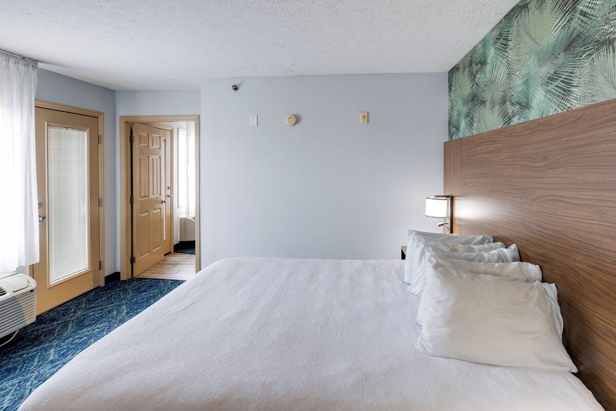 A hotel room with a large bed, a wooden headboard, a nightstand with a lamp, a window with curtains, and a door leading to another room.
