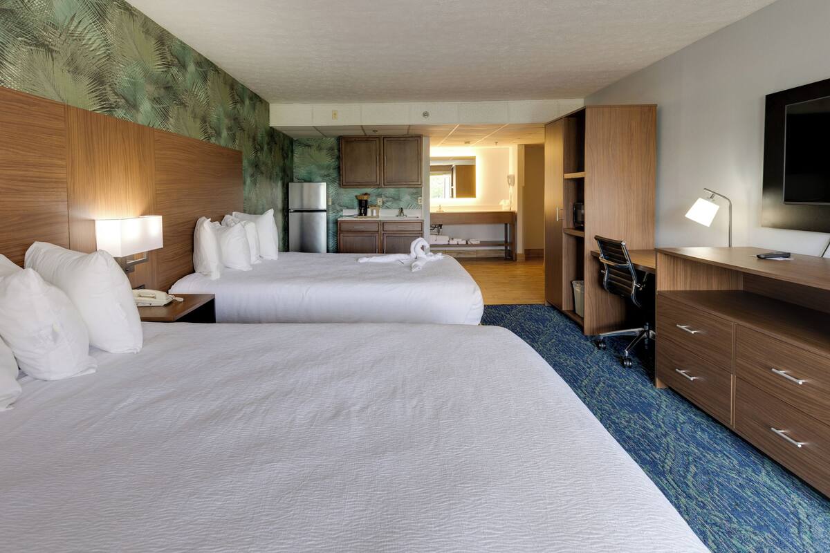 A hotel room with two beds, a kitchenette, a TV, and a desk with a chair.